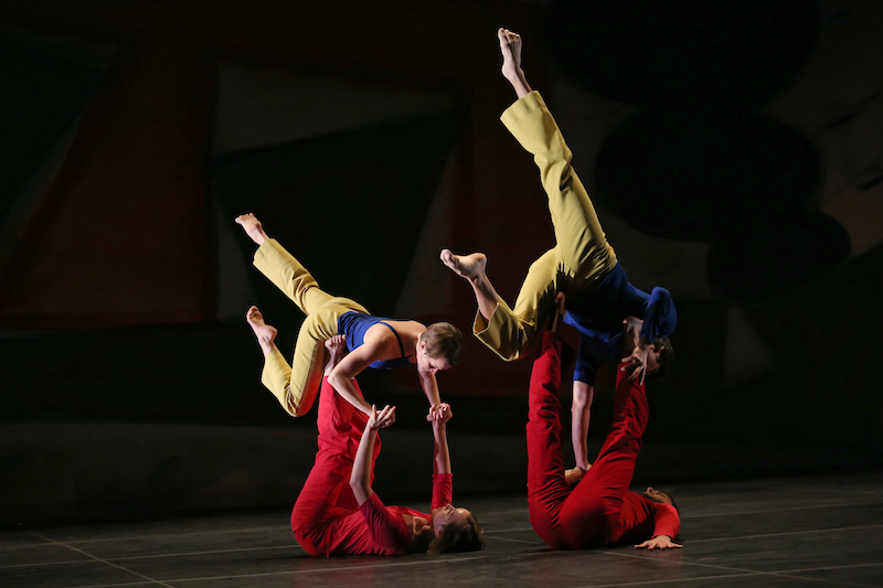 Two dancers in red on the floor hold up their respective partners by their feet and hands. It looks like their partners are floating on the stomachs in the air